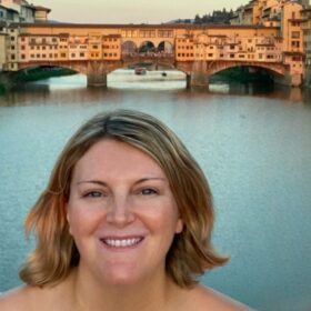 A lady with blonde hair looking at the camera with a view of Florence's Ponte Vecchio and River Arno in the background.