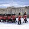 Foot Guards dressed in red and black with black bearskin hats marching in front of Buckingham Palace in the snow.