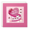 Pink square card with an illustration of a heart shaped box of chocolates with the text 'Be My Valentine' on the lid. The card features a clear window filled with confetti hearts.