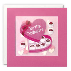 Pink square card with an illustration of a heart shaped box of chocolates with the text 'Be My Valentine' on the lid. The card features a clear window filled with confetti hearts.