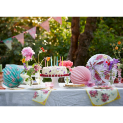 A table sits in a garden on a sunny day with bunting hanging from the trees in the background.The table is set for a party with cakes, flowers and three floral paper lantern decorations.