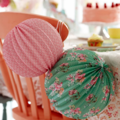 A pink and a green floral paper lantern decoration hang from the back of a chair which sits at a table upon which there are cakes and treats.