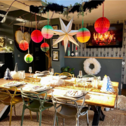 A dinner table is set for a party. Hanging from the ceiling are bright, colourful paper honeycomb decorations in ombre designs.