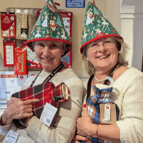 Two ladies stand in a Cards for Good Causes shop wearing festive tea cosies on their heads and holding checked fleece hot water bottles.