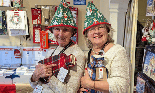 Two ladies stand in a Cards for Good Causes shop wearing festive tea cosies on their heads and holding checked fleece hot water bottles.