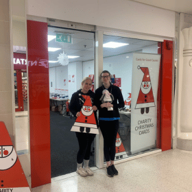 Two ladies stand outside a Cards for Good Causes Christmas shop. They are holding a cardboard Santa and a cuddly penguin toy.