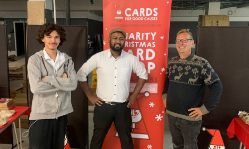 Three men stand in front of a Cards for Good Causes pop up banner in a charity Christmas shop.