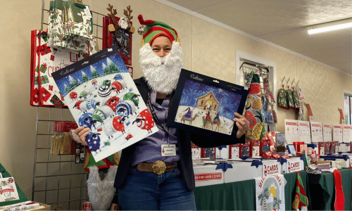 A lady wearing a Christmas elf hat and beard. She is holding two advent calendars and is standing in a Cards for Good Causes pop up Christmas shop.