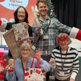 Four people wearing Christmas accessories and holding Christmas gifts in the Christmas shop at The Forum, Norwich.