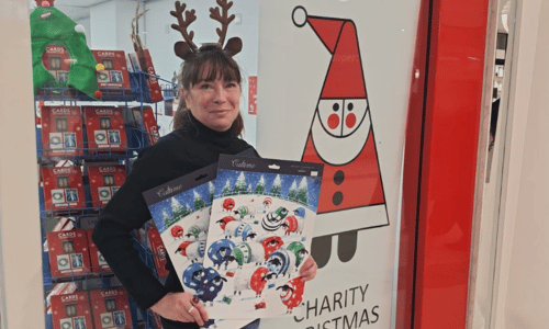A lady is wearing a reindeer antler headband and holding two advent calendars. She is standing outside a Cards for Good Causes pop up Christmas shop.