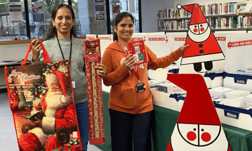 Two ladies hold up Christmas wrapping paper, bags and a cardboard Santa in a Cards for Good Causes shop.