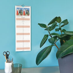 Wall-mounted calendar with illustrated header, placed beside a leafy potted plant, white pen holder with scissors, and green glass on a pastel blue surface.