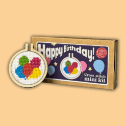 Cross stitch mini kit box featuring five colourful balloons in a small embroidery hoop, with festive packaging including stars and balloon illustrations. Large white text reads “Happy Birthday!” and notes that a keyring is included.