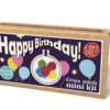 Cross stitch mini kit box featuring a colourful balloon design in a small embroidery hoop, with bright packaging and large white text reading “Happy Birthday!” Includes note that a keyring is included.