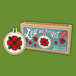 Cross stitch mini kit box featuring a red poppy with green leaves in a small embroidery hoop, set against a green background with illustrated poppies on the box. Includes cursive text “Lest we forget” and notes that a keyring is included.
