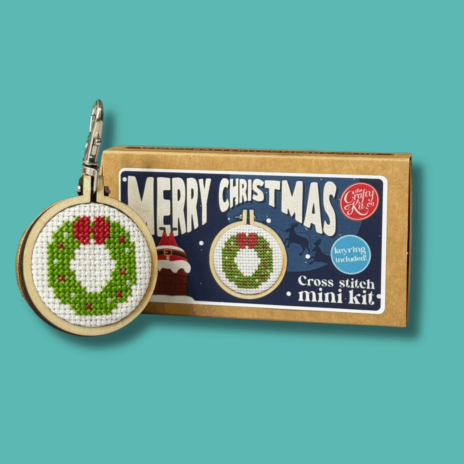 Cross stitch mini kit box featuring a festive wreath design in a small embroidery hoop, set against a green background. Includes “Merry Christmas” text on the box. Next to the box is a finished wreath embroidery hoop