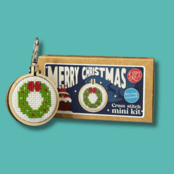 Cross stitch mini kit box featuring a festive wreath design in a small embroidery hoop, set against a green background. Includes “Merry Christmas” text on the box. Next to the box is a finished wreath embroidery hoop