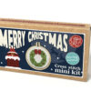 Cross stitch mini kit box with bold “Merry Christmas” text, festive wreath design in a small embroidery hoop, and cartoon chimney with an upside down Santa who's legs are sticking out.
