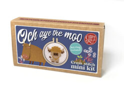 Cross stitch mini kit box featuring a Highland cow design in a small embroidery hoop. A cartoon cow is featured on the box, with floral accents and playful blue background. Includes text “Och aye the moo” and notes that a keyring is included.