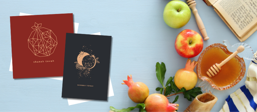 A pale blue background with two Shanah Tovah Greetings Cards next to apples, pomegranates, a jar of honey.