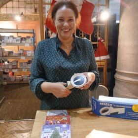 A lady holds a Braille labelling machine. There are Christmas cards on a table in front of her and stockings and Santa hats on the wall behind her.