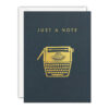 A dark blue-grey card with an illustration of a typewriter in gold foil. Text above reads 'Just a Note' in gold foil.