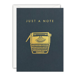 A dark blue-grey card with an illustration of a typewriter in gold foil. Text above reads 'Just a Note' in gold foil.
