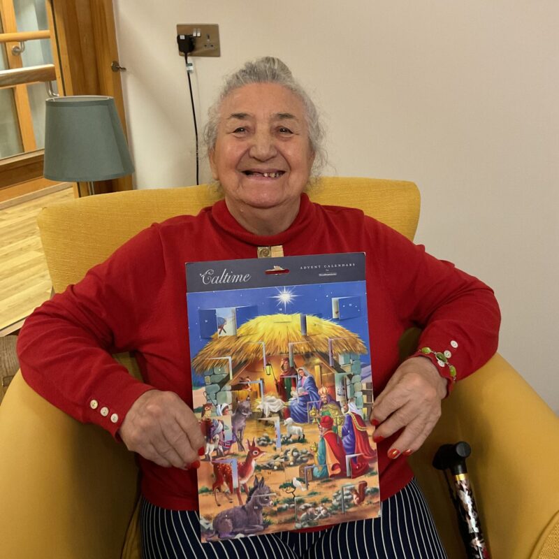 A lady wearing a red jumper smiles as she holds a large nativity advent calendar.