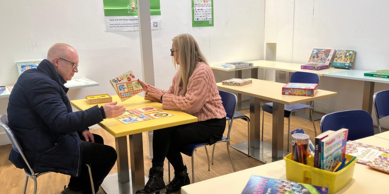 A man and woman sit at a table playing a board game. There are other tables, jigsaws and games around them.