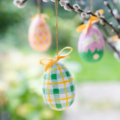 Three decorative Easter eggs hanging from a pussy willow branch, including a green‑and‑yellow check egg in the foreground and two patterned eggs in pink tones behind, all tied with yellow ribbon bows against a blurred garden background.