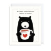 A white card with an illustration of a black bear holding a large white mug with a red heart on the front. Text reads 'Happy Birthday. Young at Heart'.