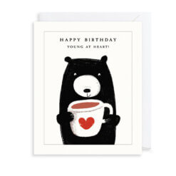 A white card with an illustration of a black bear holding a large white mug with a red heart on the front. Text reads 'Happy Birthday. Young at Heart'.