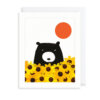 A white card with an illustration of a black bear peeking out from yellow sunflowers. There is a round orange sun above the bear.