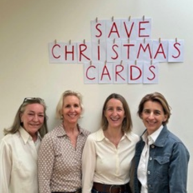 Four ladies stand together, smiling at the camera. Behind them on the wall are bits of paper with letters which spell out 'Save Christmas Cards'.